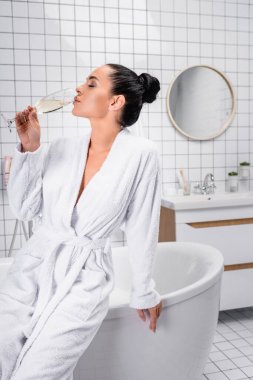 Brunette woman drinking champagne near bathtub at home  clipart