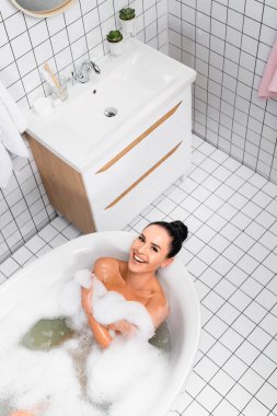 Top view of cheerful woman smiling at camera while lying in bathtub with lather  clipart
