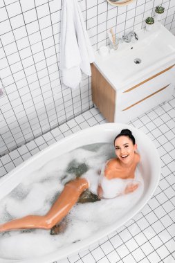Modern banyoda köpükle banyo yaparken kameraya gülümseyen esmer kadının en iyi görüntüsü. 