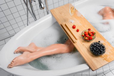 Şampanya bardağının yanında köpükle banyo yapan bir kadın ve tepside taze böğürtlenler. 