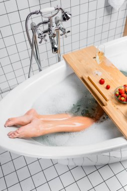Cropped view of glass of champagne near strawberries on tray and legs of woman in bathtub with foam  clipart