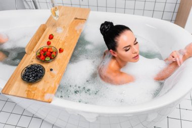 Esmer kadın dutların yanında köpükle banyo yapıyor ve şampanya da ahşap tepside. 
