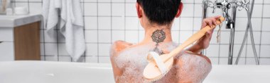 Back view of tattooed woman using brush in bathtub with soapsuds, banner  clipart