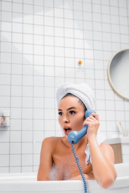 Kafasında havlu olan heyecanlı bir kadın küvette telefonla konuşuyor. 