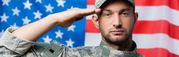 patriotic military man in uniform and cap giving salute near american flag on blurred background, banner