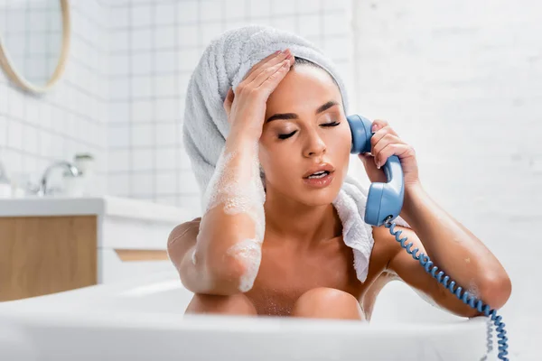 Exhausted woman in foam and towel on head talking on telephone while taking bath