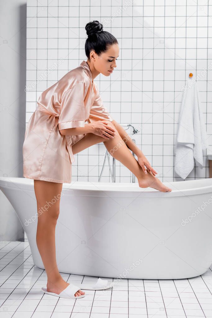 Side view of brunette woman in satin bathrobe touching leg near bathtub in modern bathroom 