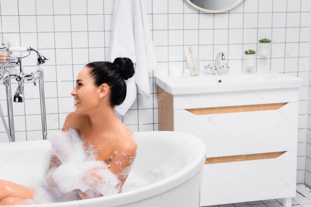 Brunette woman smiling away while sitting in bathtub with foam 