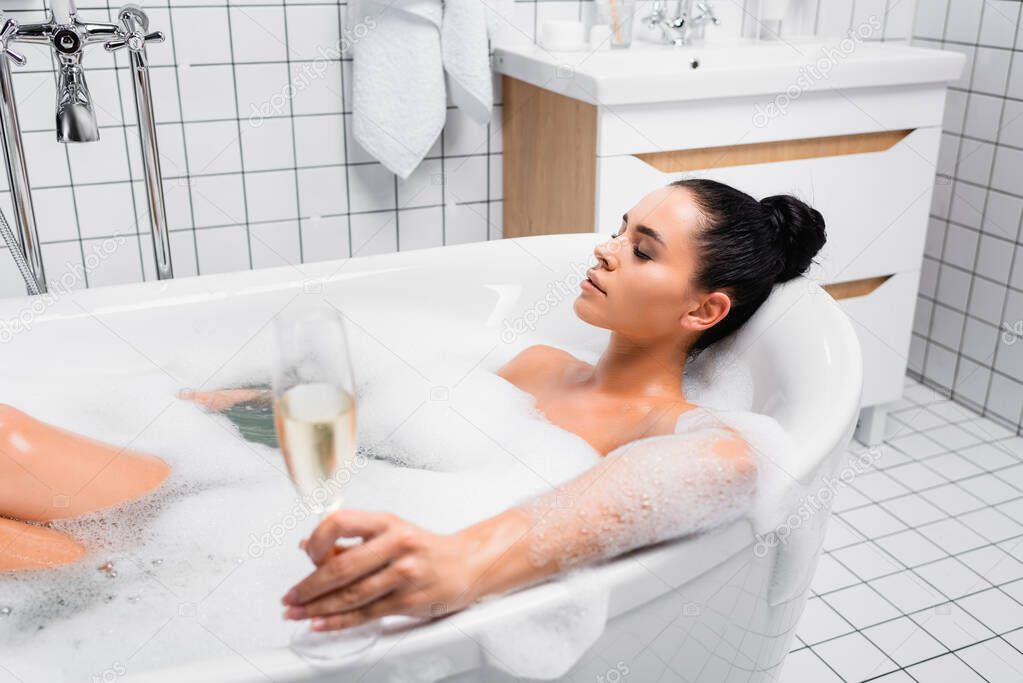 Young woman with closed eyes holding glass of champagne on blurred foreground while taking bath 
