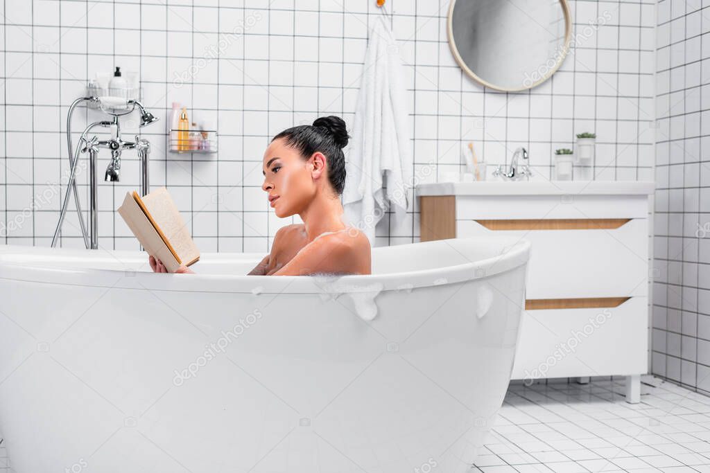 Brunette woman reading book while relaxing in bath with soapsuds at home 