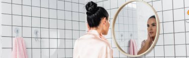 Young woman touching neck near mirror in bathroom, banner  clipart