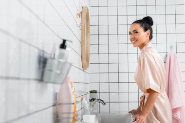 Lavabonun yanındaki kameraya gülümseyen genç bir kadının yan görüntüsü ve banyodaki bulanık ön plandaki tuvalet malzemeleri. 