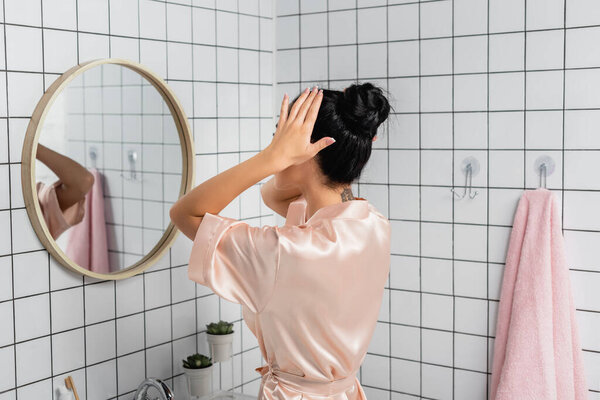 Young woman in satin bathrobe touching hair near mirror in bathroom 