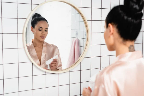 Young woman holding tube with cosmetic cream near mirror in bathroom