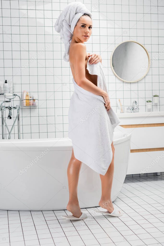 Young woman in towels looking at camera near bathtub at home 