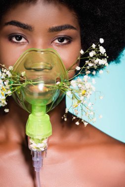 Solunum cihazında çiçekleri olan Afro-Amerikalı genç bir kadın mavi arka planda izole edilmiş.