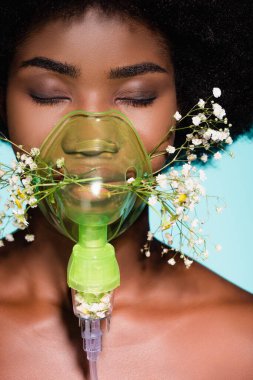 Solunum cihazında çiçekleri olan Afro-Amerikalı genç bir kadın mavi arka planda izole edilmiş.