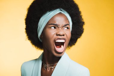 Kızgın, Afro-Amerikalı, mavi kıyafetli genç bir kadın sarı renkte izole bir şekilde bağırıyor. 