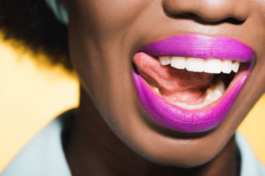 cropped view of african american young woman with purple lips showing tongue isolated on yellow  clipart