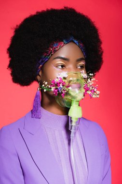 Afro-amerikan genç bayan mor renkli şık kıyafetli, solunum cihazında çiçekli, kırmızıda izole edilmiş.
