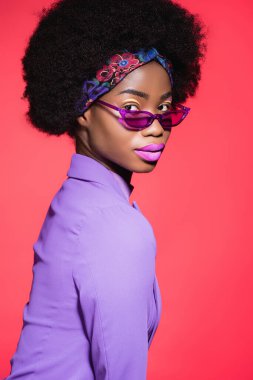 Kırmızıda izole edilmiş mor giysili ve güneş gözlüklü Afro-Amerikan genç bayanın yan görüntüsü