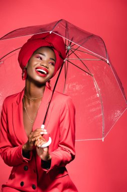 happy african american young woman in stylish outfit and turban holding umbrella isolated on red clipart