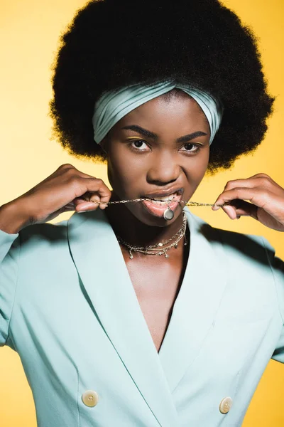 身穿蓝色时髦服装 嘴里衔着项链 与黄色隔离的非洲裔美国年轻女子 — 图库照片