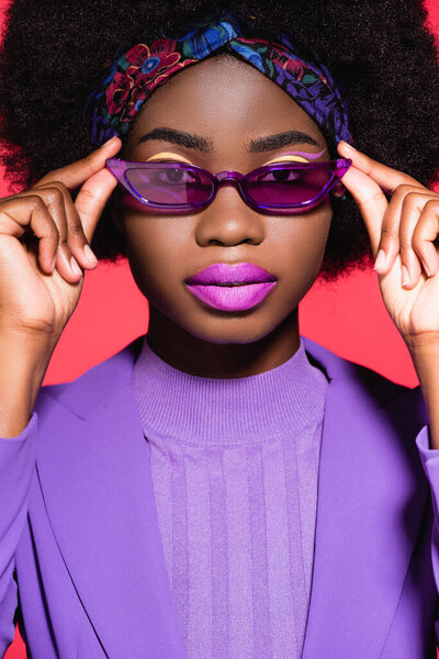 Африканская американская молодая женщина в фиолетовом стильном наряде и солнечных очках, изолированных на красном