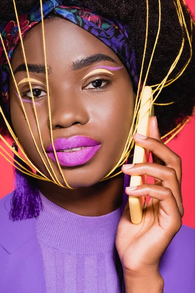 african american young woman in purple stylish outfit with yellow strings on face holding smartphone isolated on red