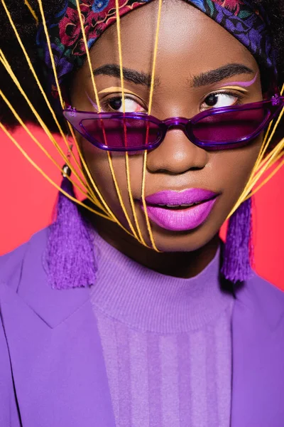 african american young woman in purple stylish outfit and sunglasses with yellow strings on face isolated on red