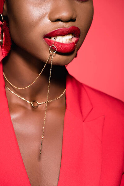 cropped view of african american young woman in stylish outfit holding necklace in mouth isolated on red