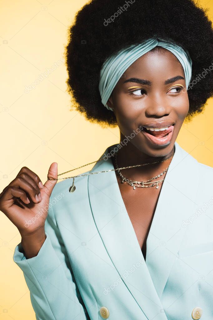 african american young woman in blue stylish outfit showing necklace isolated on yellow