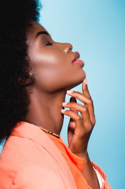 Yandan bakınca turuncu kıyafetli, çenesine dokunan mavi tenli Afro-Amerikalı genç bir kadın görülüyor.