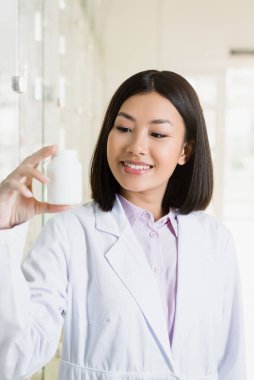 Beyaz önlüklü neşeli Asyalı eczacı ilaç şişesine bakıyor. 