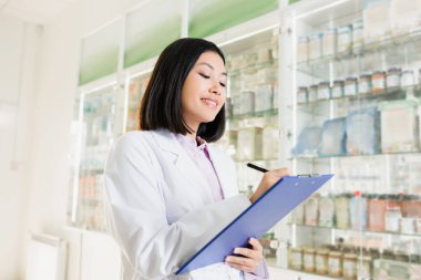 cheerful asian pharmacist in white coat holding pen near clipboard in drugstore  clipart