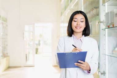 smiling asian pharmacist in white coat writing on clipboard in drugstore  clipart