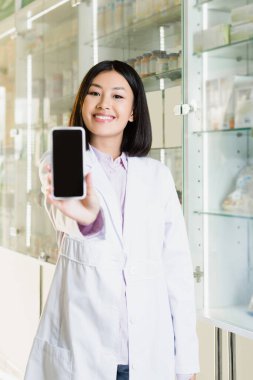 cheerful asian pharmacist in white coat holding smartphone with blank screen on blurred foreground  clipart