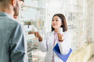 asian pharmacist in white coat holding clipboard and bottles with medication near man in drugstore  clipart