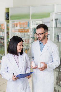 Eczanedeki Asyalı meslektaşının yanında elinde ilaç şişeleri tutan beyaz önlüklü mutlu bir eczacı.