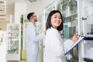 happy asian pharmacist in white coat writing on clipboard near colleague on blurred background clipart