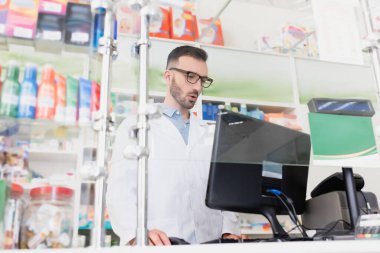 surprised pharmacist in white coat and eyeglasses looking at computer monitor in drugstore clipart
