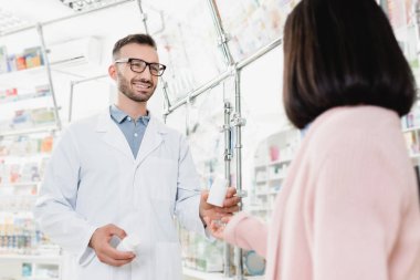 Gözlüklü ve beyaz önlüklü neşeli eczacı müşterinin yakınındaki bulanık ön planda hapları tutan beyaz önlüklü.