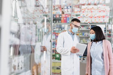 Tıbbi maskeli eczacı eczanedeki Asyalı kadının yanında dijital tablet tutuyor.