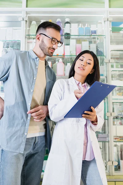 Morena Asiático Farmacéutico Blanco Abrigo Celebración Portapapeles Cerca Barbudo Cliente — Foto de Stock