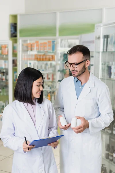 在药店里 穿着白衣的快乐药剂师拿着装有药物的瓶子 和亚洲同事在一起 — 图库照片