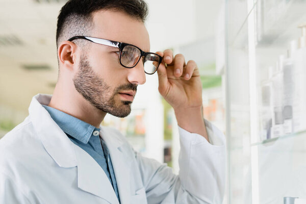 bearded pharmacist in white coat adjusting eyeglasses in drugstore 