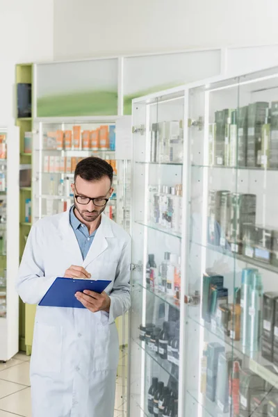 Szakállas Gyógyszerész Szemüvegben Írás Vágólapra Miközben Gyógyszertárban Ellenőrzi Gyógyszereket — Stock Fotó