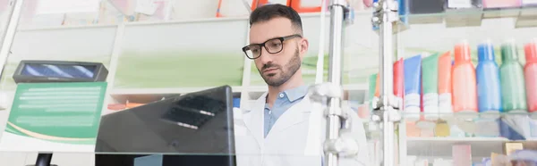 pharmacist in white coat and eyeglasses looking at computer monitor in drugstore, banner