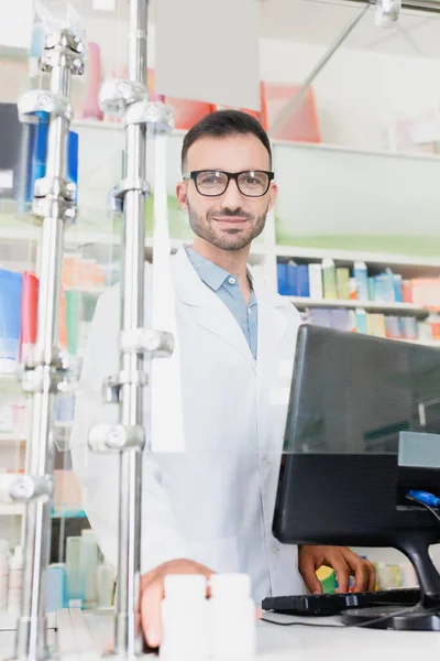 药店里身穿白衣 戴着眼镜 微笑的药剂师 在电脑监视器前看着摄像机 — 图库照片