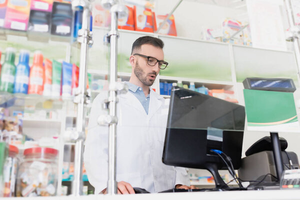 surprised pharmacist in white coat and eyeglasses looking at computer monitor in drugstore